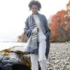 Swans Island's Penobscot Throw is woven in Maine with organic Merino wool and organic American Cotton. Shown here in Graphite with White on model at rocky beach with fall foliage in the background.