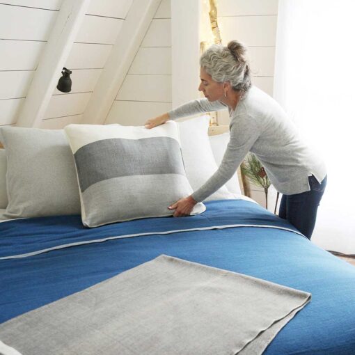 Swans Island Company's Maine Coast Pillows. Handwoven in Maine with soft hand-dyed organic merino wool. Shown in Charcoal. Woman placing pillow on bed with indigo blue blanket.