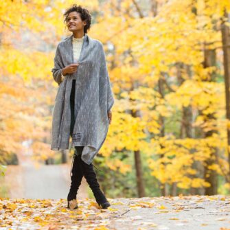 Swans Island Handwoven Firefly Wrap. Made in Maine with hand-dyed merino wool / silk yarn. Shown here in Onyx. Woman is walking on a road in the woods surrounded by golden fall foliage.
