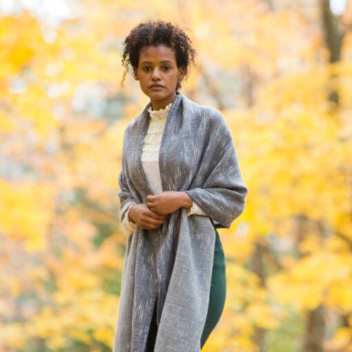 Swans Island Handwoven Firefly Wrap. Made in Maine with hand-dyed merino wool / silk yarn. Shown here in Onyx. Woman is walking on a road in the woods surrounded by golden fall foliage.