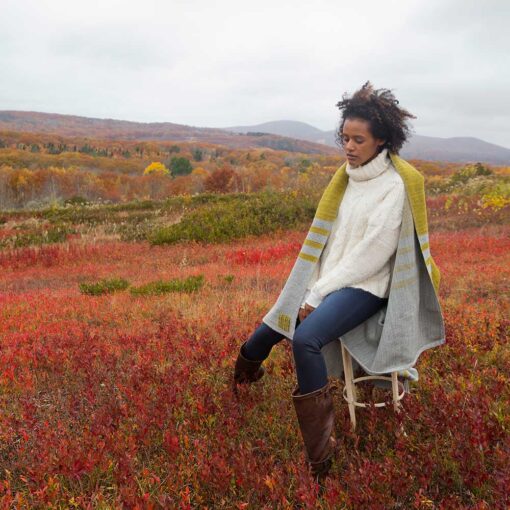 Swans-Island Mt. Kineo-Throw shown here in Granite + Lichen, hand-dyed and handwoven in Maine. Woman wrapped in throw in a colorful red Maine autumn blueberry field with all foliage in the background.