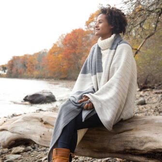 Swans Island Company's Rangeley Throw captures the essence of the Maine Coast in shades of grey. Woven in Maine with ultra-soft organic merino wool and cotton. Hand-dyed. Shown here in Graphite + Pewter.. Woman sitting on driftwood log on a rocky shore wrapped in throw, with fall foliage in the background.