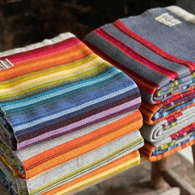 Swans Island Company's Limited Edition Prism Series Throws. Nine rainbow-inspired throws designed by our team of talented weavers. Each throw in this limited edition drop is unique! A stack of beautiful finished Prism Throws await their new homes.