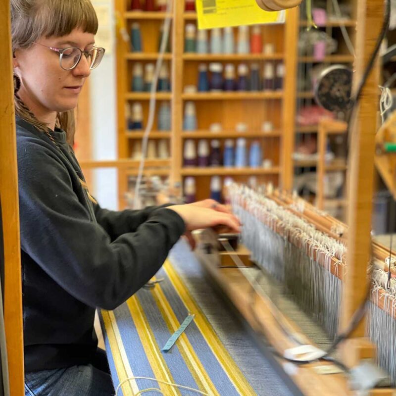 Swans Island Company's Limited Edition Prism Series. Nine rainbow-inspired throws designed by our team of talented weavers. Each throw in this limited edition drop is unique! Here's Chloe working at the loom making one of the nine unique throws.