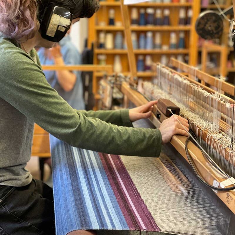 Swans Island Company's Limited Edition Prism Series. Nine rainbow-inspired throws designed by our team of talented weavers. Each throw in this limited edition drop is unique! Here's Riley working at the loom making one of the nine unique throws.